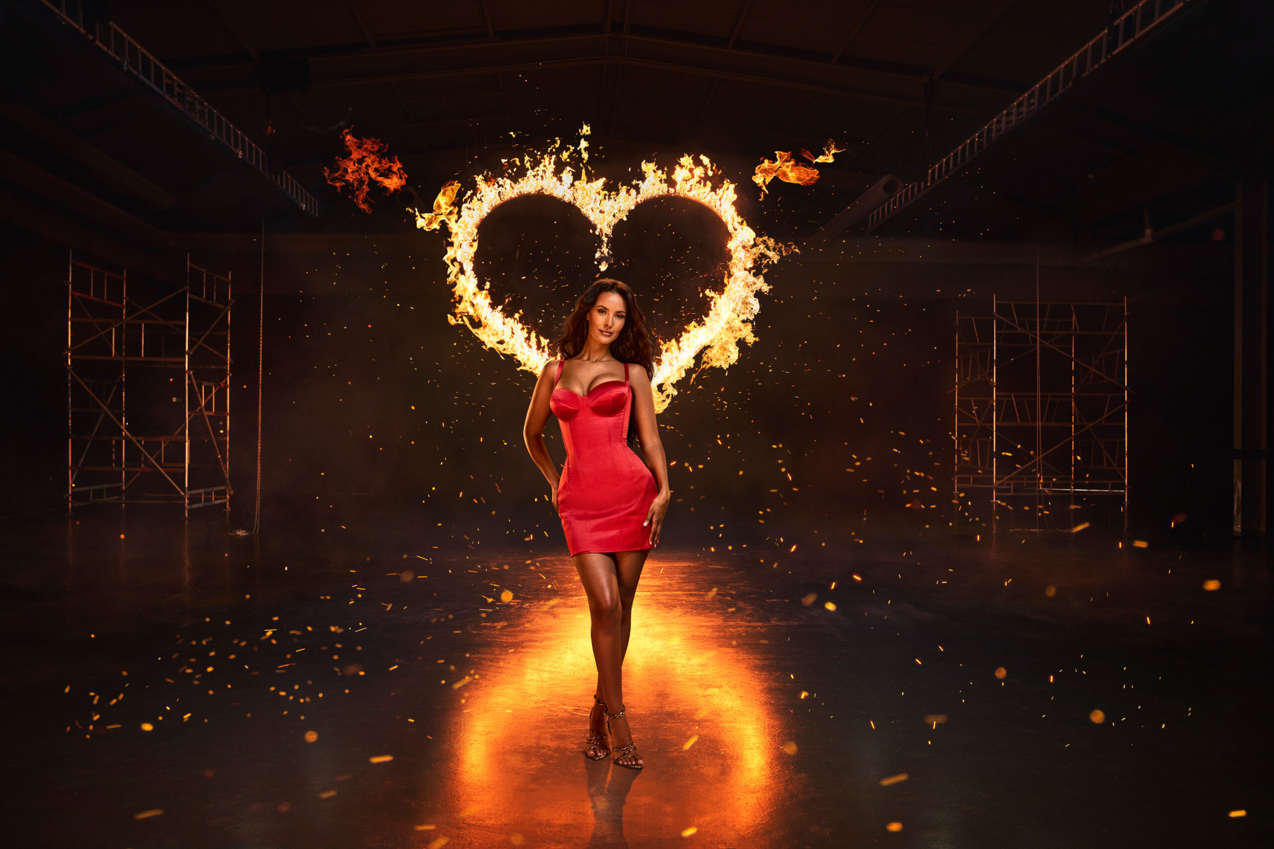 Love Island Series 11 shot by Ian Hippolyte. In the photograph, a mixed-race woman, Maya Jama, stands wearing a red cocktail dress, in a dark warehouse location. Behind her is flames in the shape of a love heart.